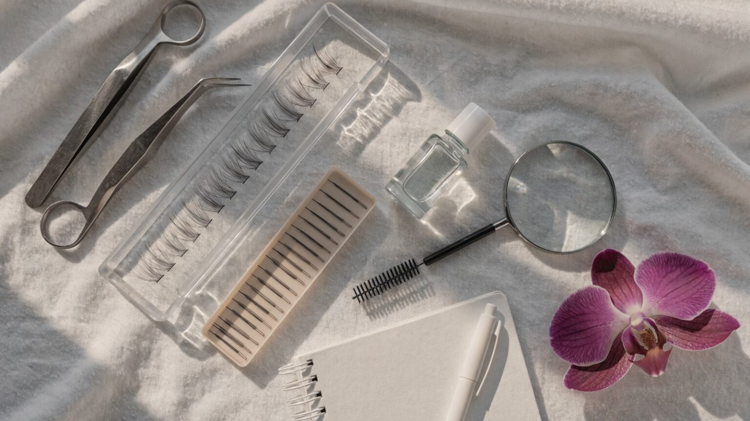 Helles Flatlay mit Wimpernpinzetten, Wimpernfächern, Wimpernkleber-Entferner, Spoolie, Lupe und plum-farbener Orchidee für die Übernahme von Fremdarbeit