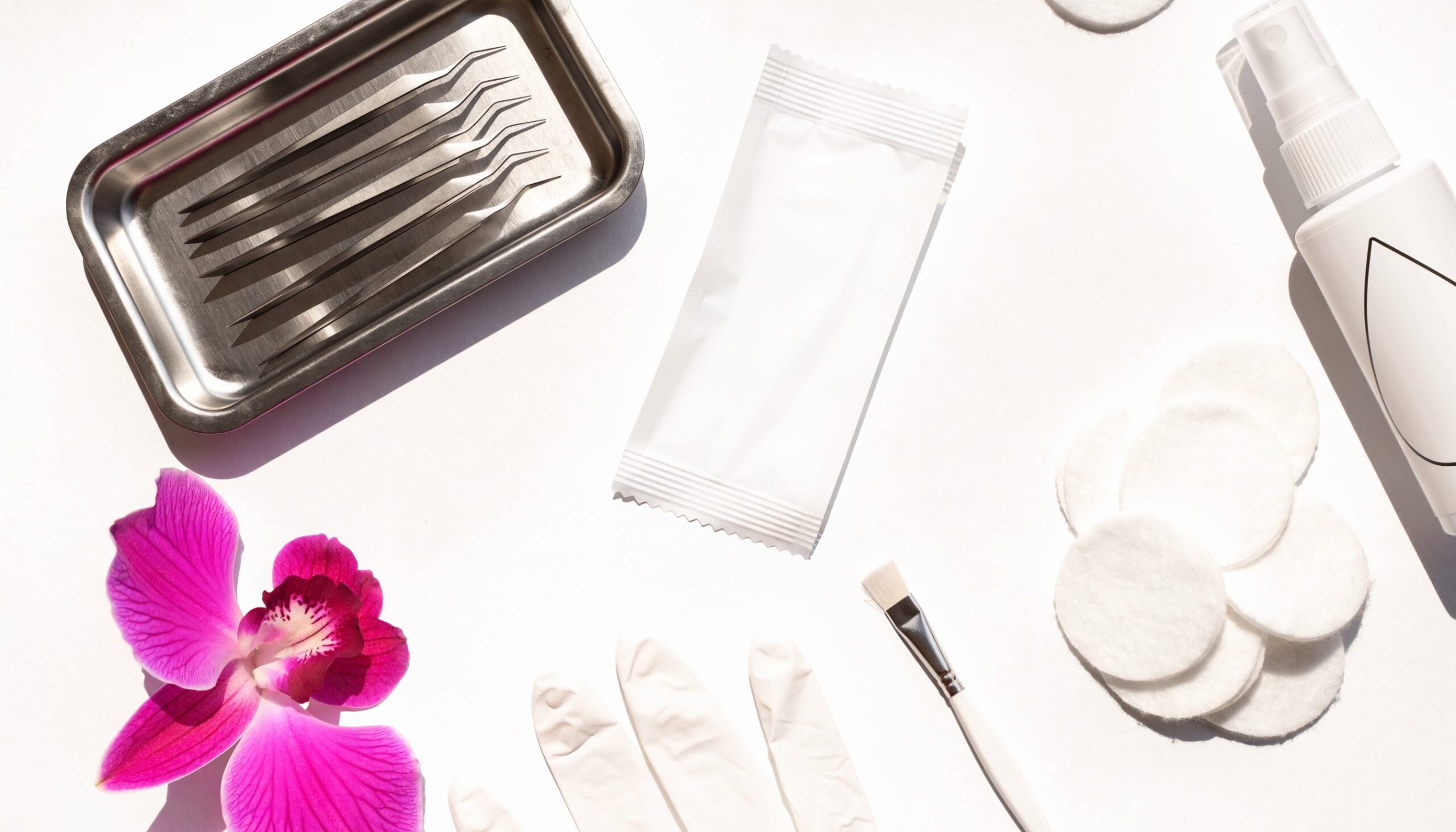 Helles weißes Hygiene-Flatlay: Lash-Pinzetten auf Sterilisationstablett, Steri-Beutel, Desinfektionsspray, Handschuh und Wattepads mit Magenta-Orchidee