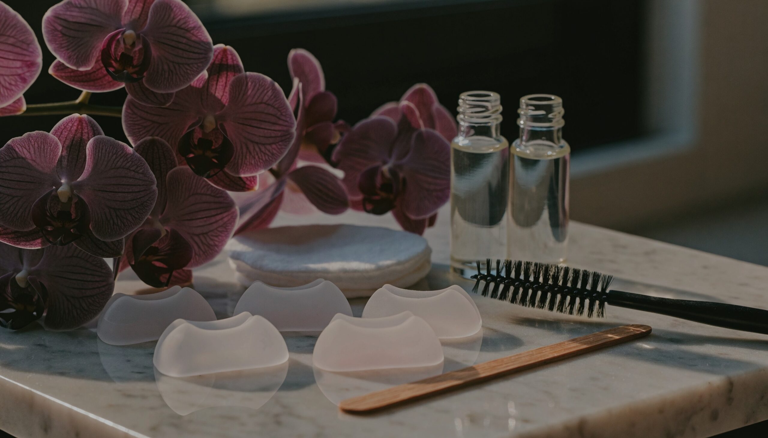 Wimpernlifting Wien: Silikonpads in verschiedenen Größen, Wimpernbürstchen, Applikator und Glasfläschchen auf einem hellen Marmortisch im Studio