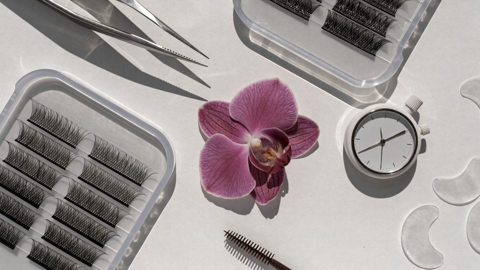 Helles Flatlay mit Wimpernpinzette, Wimpernfächern, Timer, Spoolie, Pads und plum-farbener Orchidee zum Thema Dauer eines Wimpern-Neusets