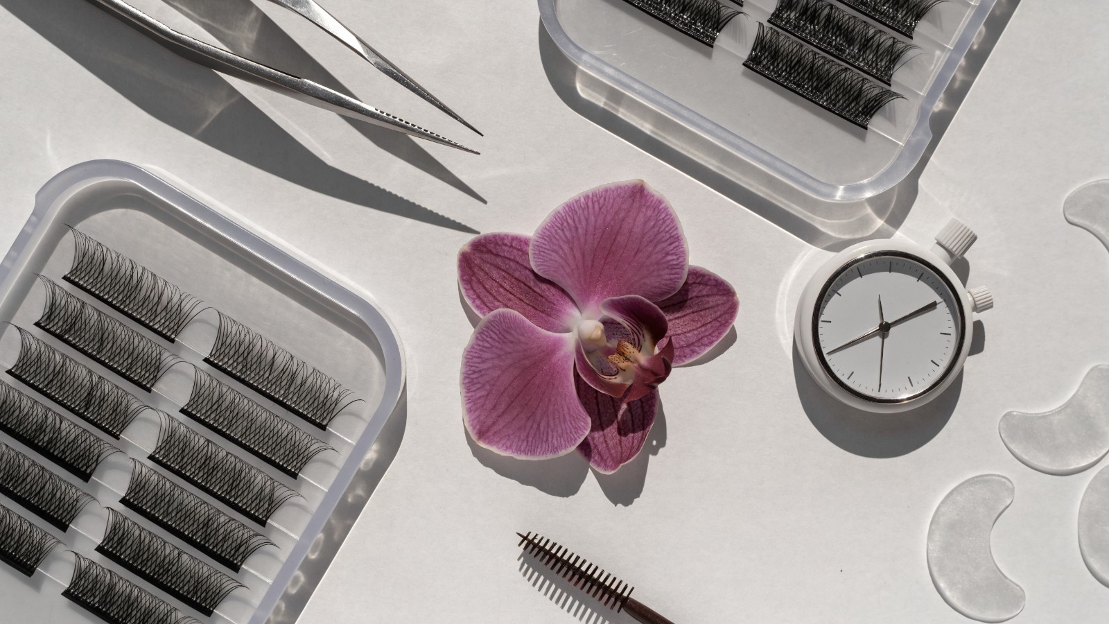 Helles Flatlay mit Wimpernpinzette, Wimpernfächern, Timer, Spoolie, Pads und plum-farbener Orchidee zum Thema Dauer eines Wimpern-Neusets