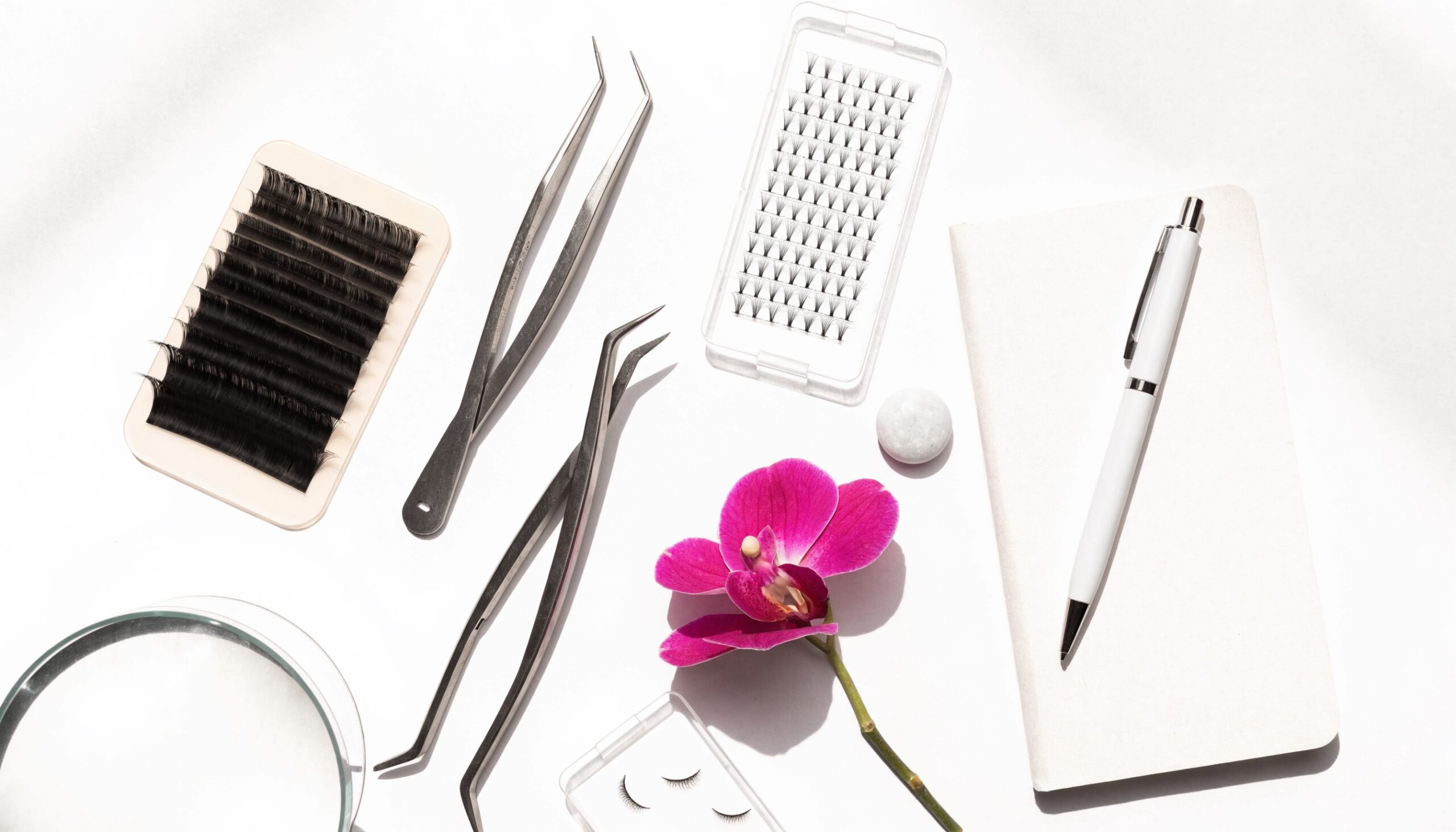 Helles weißes Flatlay für die Wimpernvolumen-Schulung: Lash-Tray, Volumen-Fächer, Pinzetten, Notizbuch und Magenta-Orchidee